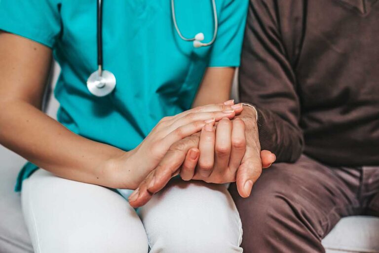 An older adult sitting with a caregiver, checking medication and talking in a comfortable residential setting.